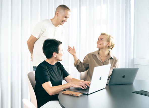 Kolme kollegaa keskustelemassa työasioista pöydän ääressä kannettavien tietokoneiden kanssa valoisassa toimistossa.